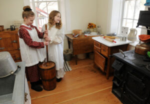 Children dressed in period clothing making butter in the kitchen of the Bown House