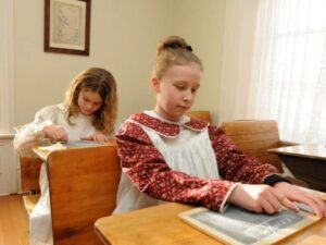 Children dressed in period clothing writing on chalkboards in the schoolroom at the Bown House