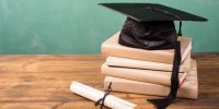 Spring graduation time.  A black cap/motarboard with black tassel lies on top of a stack of textbooks.  A diploma in front.  All objects lie on a rustic wooden desk with green chalkboard in background.  Copyspace to side.  No people.  Education themes.