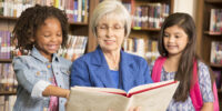 Teacher, librarian reads book to elementary students in library or classroom. Senior adult teacher or librarian reads a book to elementary students in school classroom or library setting. She is helping the girls learn to read or learn English.
