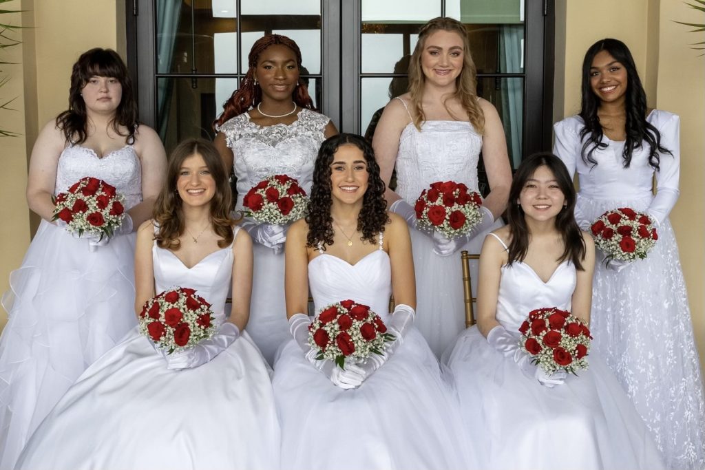 Seven high school senior girls wearing white gowns, holding red flowers