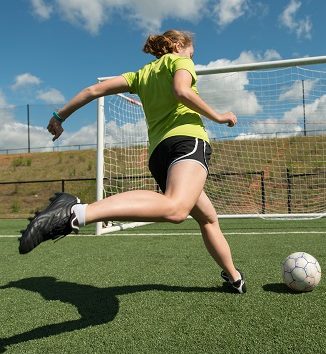 Young adult female soccer player practices shots on goal on a beautiful turf field