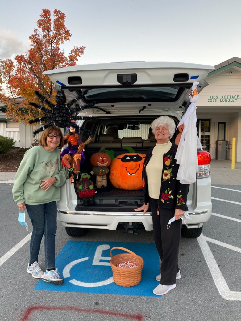 A picture containing sky, outdoor, road, 2 women handing out candy from the trunk of a car